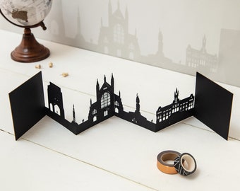 May include: A black paper skyline silhouette with a zigzag design, featuring architectural details. A globe and decorative tape rolls are in the background. The skyline is displayed on a white surface.
