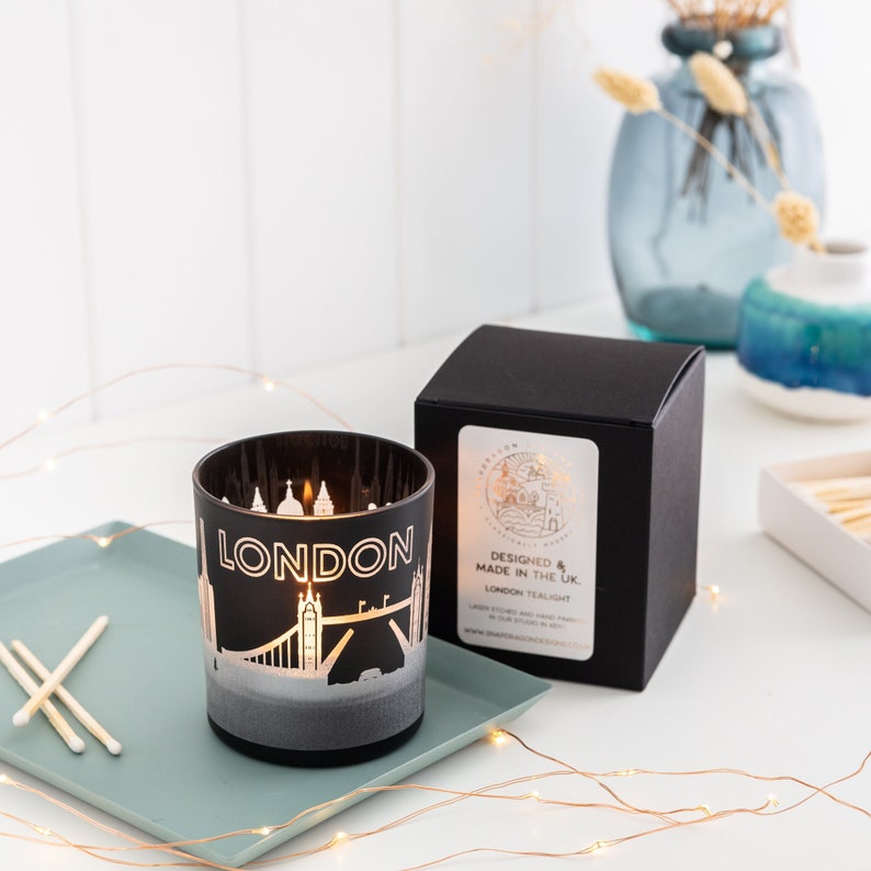 May include: A black London-themed candle in a glass holder, featuring a cityscape design with the word "LONDON". The candle sits on a teal tray with matches, next to a black box with a gold label. Decorative lights and vases are in the background.