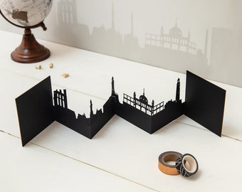 May include: Black paper cityscape card with a folded design. The card is folded into a zig-zag pattern and features a silhouette of a city skyline.