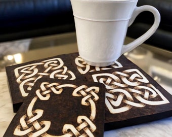 May include: Four wooden coasters with a Celtic knot design burned into the surface. The coasters are dark brown with a light brown knot design. A white ceramic mug is sitting on top of the coasters.