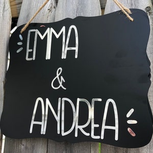 Puede incluir: Letrero negro de pizarra con una inscripci&oacute;n blanca en cursiva que dice "Emma & Andrea". El letrero tiene un borde decorativo festoneado y est&aacute; colgado de una cuerda.