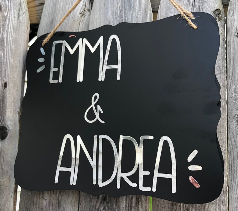 Puede incluir: Letrero negro de pizarra con una inscripci&oacute;n blanca en cursiva que dice "Emma & Andrea". El letrero tiene un borde decorativo festoneado y est&aacute; colgado de una cuerda.