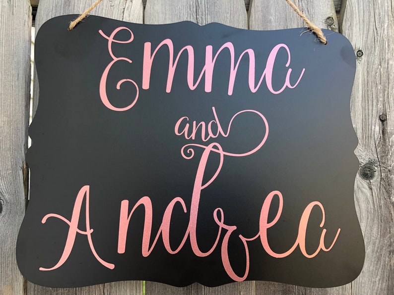 Puede incluir: Se&ntilde;al negra de pizarra con marco blanco y una inscripci&oacute;n cursiva rosa que dice "Emma y Andrea".