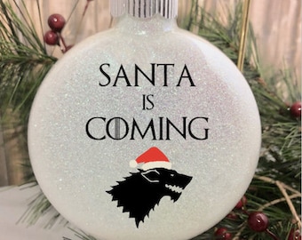 Puede incluir: Adorno navideño blanco con purpurina con el texto "Santa is Coming" y una silueta negra de un lobo huargo con un gorro de Papá Noel.