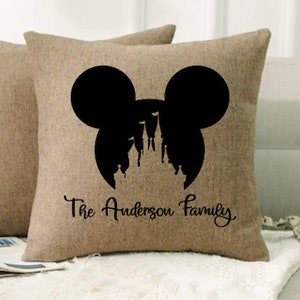 May include: A brown burlap pillow with a black silhouette of Mickey Mouse's head with Cinderella's castle inside. The text "The Anderson Family" is printed below the silhouette.