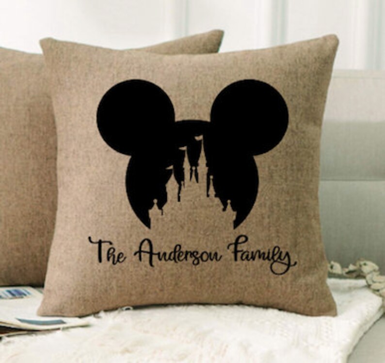 May include: A brown burlap pillow with a black silhouette of Mickey Mouse's head with Cinderella's castle inside. The text "The Anderson Family" is printed below the silhouette.