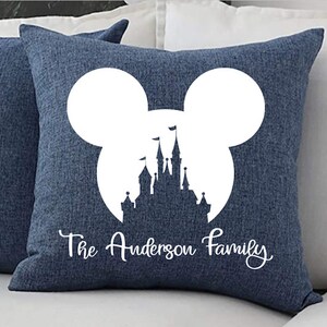 May include: A blue throw pillow with a white silhouette of Mickey Mouse's head with Cinderella's castle inside. The text "The Anderson Family" is printed below the silhouette.