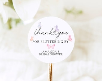May include: Round white thank you sticker with a pink and purple butterfly design. The sticker says "Thank you for fluttering by! Amanda's Bridal Shower".