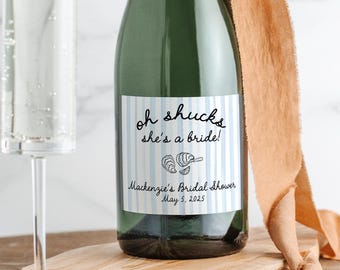 May include: A champagne bottle with a blue and white striped label that says "oh shucks she's a bride!"  Mackenzie's Bridal Shower May 5, 2025. The bottle is tied with a brown ribbon and a champagne flute is next to it.