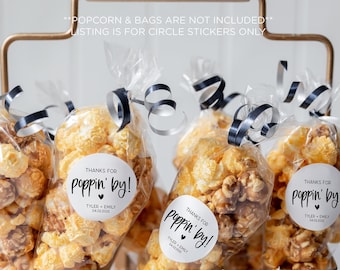 May include: A gold metal stand filled with clear bags of popcorn. Each bag has a white circle sticker with black text that says "Thanks for poppin' by!" with a small heart symbol.