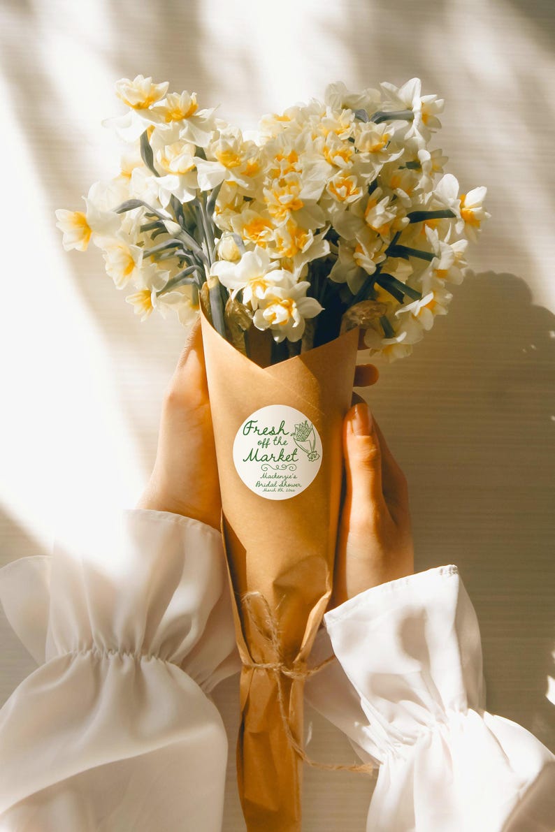 May include: A bouquet of white and yellow daffodils wrapped in brown paper with a white circle sticker that says "Fresh off the Market" and "Mackenzie's Bridal Shower" in green text.