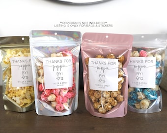 May include: Four clear plastic bags with zipper closures filled with different colored popcorn. Each bag has a white sticker with the text "Thanks for poppin' by!" and the names "Tyler & Emily" and the date "05.01.22".