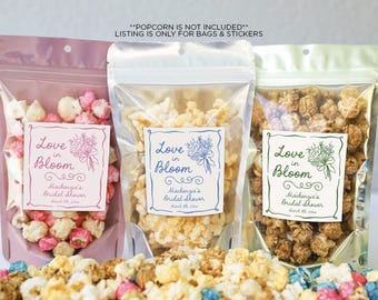 May include: Three clear plastic bags filled with popcorn, each with a label that says "Love in Bloom" and includes the name "Mackenzie's Bridal Shower". The labels are pink, blue, and green.