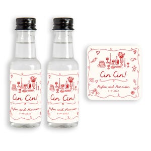 May include: Two small clear glass bottles with black caps and labels featuring red illustrations and text. The labels read "Cin Cin!" and include names and a date. A matching coaster is also present.
