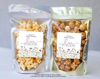 May include: Two clear plastic bags filled with popcorn. Each bag has a white square label with the text "Ashley's Bridal Shower Love in Bloom" and a floral design.