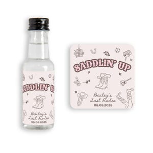 May include: A pink and white mini liquor bottle with a label that says "Saddlin' Up" and "Bailey's Last Rodeo 05.05.2025". The label has a western theme with illustrations of cowboy boots, hats, and musical notes.