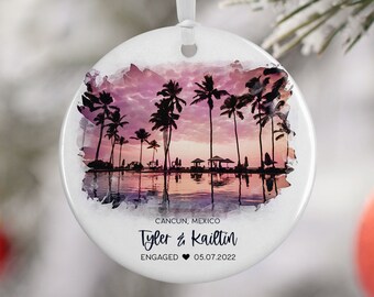 May include: A white round ornament with a watercolor painting of a sunset over a beach with palm trees. The text "CANCUN, MEXICO Tyler & Kaitlin ENGAGED 05.07.2022" is printed on the ornament.