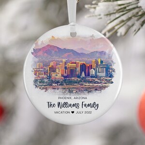 May include: A white round Christmas ornament with a watercolor painting of the Phoenix skyline. The text "PHOENIX, ARIZONA The Williams Family VACATION JULY 2022" is printed on the ornament.