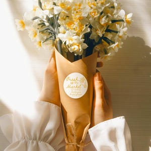 May include: A bouquet of white and yellow daffodils wrapped in brown paper with a white sticker that says "Fresh off the Market" and "Madison's Bridal Shower" in gold lettering.
