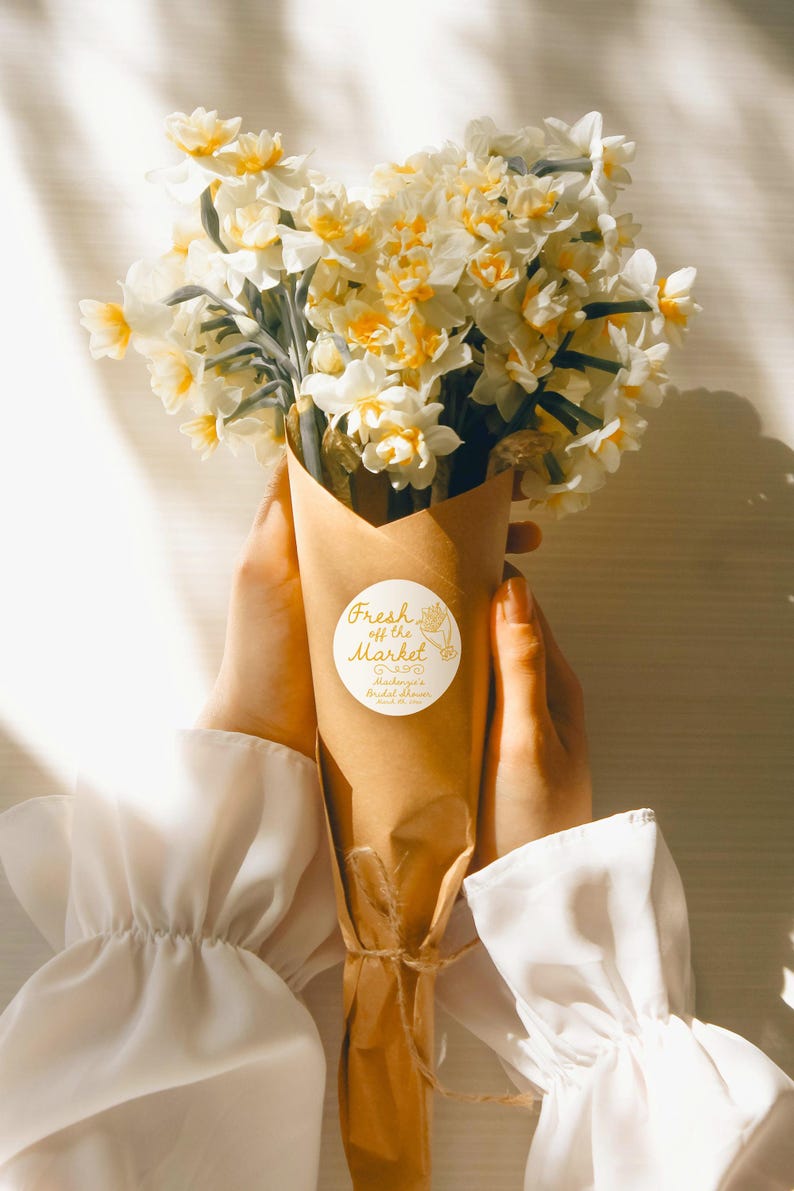 May include: A bouquet of white and yellow daffodils wrapped in brown paper with a white sticker that says "Fresh off the Market" and "Madison's Bridal Shower" in gold lettering.