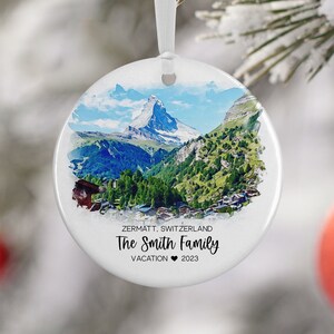 May include: White ceramic Christmas ornament with a watercolor image of the Matterhorn mountain in Zermatt, Switzerland. The text "Zermatt, Switzerland The Smith Family Vacation ♥ 2023" is printed on the ornament.