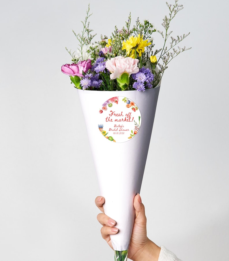 May include: A white paper cone filled with a bouquet of pink, yellow, and purple flowers. A round sticker with a watercolor floral design and the text "Fresh off the market!" is attached to the cone. The text "Bailey's Bridal Shower 04.23.2023" is also on the sticker.