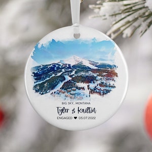 May include: A white ceramic ornament with a watercolor painting of a snowy mountain landscape. The text "Big Sky, Montana Tyler & Kaitlin ENGAGED 05.07.2022" is printed on the ornament.