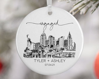 May include: White round ceramic bauble with a black and white illustration of a city skyline. The text "engaged" is written in cursive at the top of the bauble. The text "Tyler + Ashley 07.04.21" is written at the bottom of the bauble.