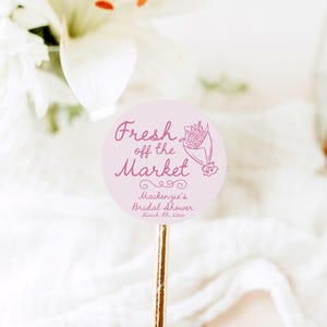 May include: Pink round sticker with the text "Fresh off the Market" in pink cursive font. A pink line drawing of a bouquet of flowers is to the right of the text. Below the text is "Mackenzie's Bridal Shower March 11, 2023" in a smaller font.