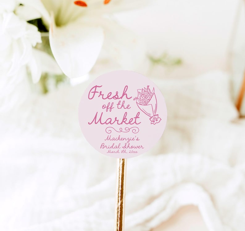 May include: Pink round sticker with the text "Fresh off the Market" in pink cursive font. A pink line drawing of a bouquet of flowers is to the right of the text. Below the text is "Mackenzie's Bridal Shower March 11, 2023" in a smaller font.