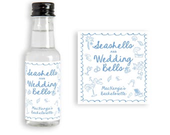 May include: A small clear glass bottle with a black cap and a white label. The label has blue text that says "Seashells and Wedding Bells" with a blue illustration of seashells, wedding rings, and other beach-themed items. The label also says "Mackenzie's Bachelorette".