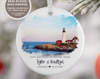 May include: A round white ceramic ornament featuring a watercolor illustration of the Portland Head Lighthouse in Maine. The text reads PORTLAND HEAD LIGHTHOUSE, MAINE, Tyler & Kaitlyn, ENGAGED, and 05.07.2022 with a heart symbol.