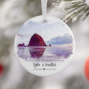 May include: A round, white, ceramic ornament with a watercolor painting of Haystack Rock at Cannon Beach, Oregon. The text "Tyler & Kaitlin ENGAGED 05.07.2022" is printed on the ornament.