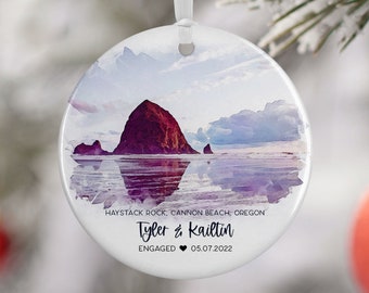 Pode incluir: Um ornamento redondo de cerâmica branca com uma pintura em aquarela de Haystack Rock em Cannon Beach, Oregon. O texto "Tyler & Kaitlin ENGAGED 05.07.2022" está impresso no ornamento.
