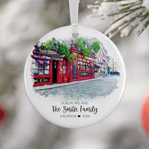 Könnte beinhalten: Ein weißer runder Weihnachtsbaumschmuck mit einem Aquarellbild eines roten Backsteingebäudes mit einem grünen Vordach und dem Text "The Temple Bar" an der Seite. Das Gebäude befindet sich auf einer Kopfsteinpflasterstraße mit anderen Gebäuden im Hintergrund. Der Text "Dublin, Irland The Smith Family Vacation 2024" ist auf den Baumschmuck gedruckt.