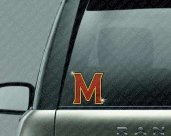 May include: A red and gold rhinestone letter "M" decal, affixed to a car window. The letter is outlined in gold and filled with red rhinestones. The background shows the car's exterior and the text "Be Dazzled Decals".