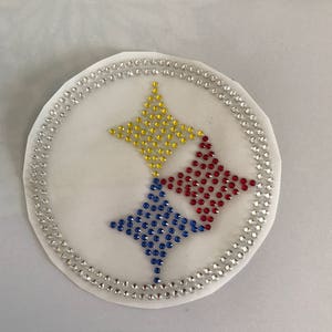 May include: A white circle with a silver rhinestone border. Inside the circle are three stars made of rhinestones. The top star is yellow, the left star is blue, and the right star is red.