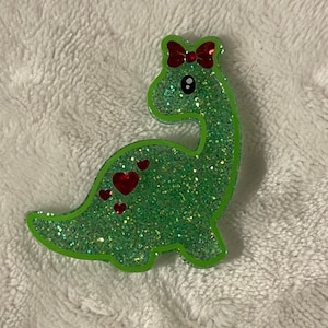 May include: A green glitter dinosaur with a red bow and red hearts on its back.