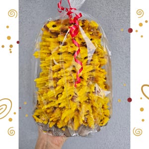 Puede incluir: Un pastel de brocheta alto y cilíndrico con una textura de color amarillo dorado, en capas y con puntas, envuelto en plástico transparente y atado en la parte superior con una cinta rizada de color rojo brillante. La superficie es irregular.