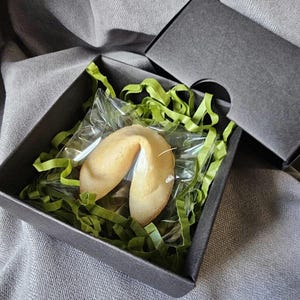 Op de afbeelding: Een fortuinkoekje in een doorzichtige plastic verpakking, genesteld in groene papiersnippers in een zwarte geschenkdoos. De doos is open en onthult de koek. De achtergrond is een grijze stof.