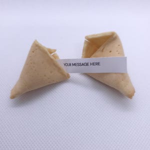 Puede incluir: Una galleta de la fortuna de color beige claro, partida en dos piezas. Una tira de papel blanco se encuentra entre las dos mitades, con el texto impreso en negro que dice YOUR MESSAGE HERE en letras may&uacute;sculas.