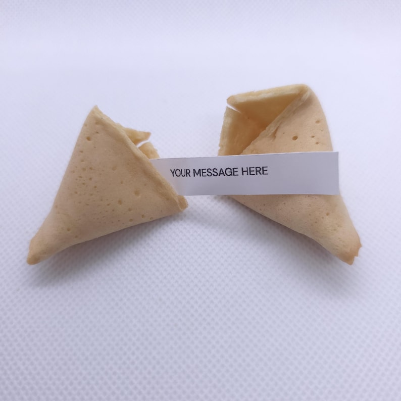 Puede incluir: Una galleta de la fortuna de color beige claro, partida en dos piezas. Una tira de papel blanco se encuentra entre las dos mitades, con el texto impreso en negro que dice YOUR MESSAGE HERE en letras may&uacute;sculas.