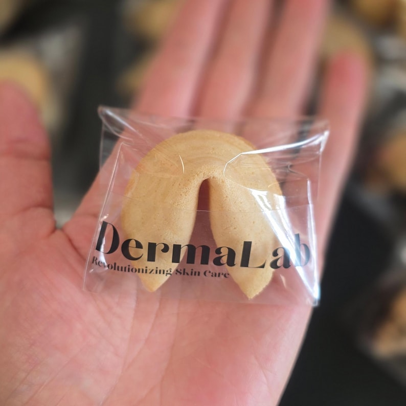Puede incluir: Una galleta de la fortuna de color tostado claro envuelta en pl&aacute;stico transparente. El envoltorio presenta el texto negro DermaLab seguido del texto m&aacute;s peque&ntilde;o Revolutionizing Skin Care impreso en el frente del film.