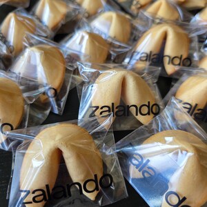 Puede incluir: Una colecci&oacute;n de galletas de la fortuna de color marr&oacute;n dorado, cada una envuelta individualmente en pl&aacute;stico transparente. La palabra zalando est&aacute; impresa en letras min&uacute;sculas negras en el frente de cada envoltorio pl&aacute;stico.