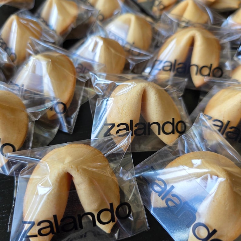 Puede incluir: Una colecci&oacute;n de galletas de la fortuna de color marr&oacute;n dorado, cada una envuelta individualmente en pl&aacute;stico transparente. La palabra zalando est&aacute; impresa en letras min&uacute;sculas negras en el frente de cada envoltorio pl&aacute;stico.