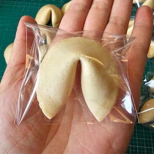 Puede incluir: Una galleta de la fortuna de color beige envuelta en una bolsa de pl&aacute;stico transparente cuadrada. La galleta tiene una forma plegada de media luna con una textura horneada suave. Los bordes son ligeramente curvos y puntiagudos.