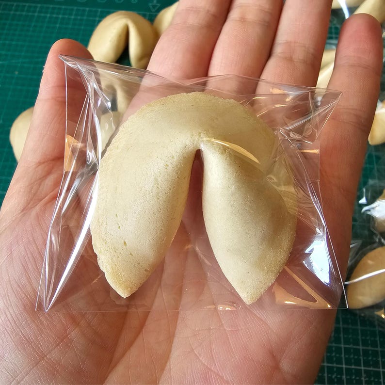Puede incluir: Una galleta de la fortuna de color beige envuelta en una bolsa de pl&aacute;stico transparente cuadrada. La galleta tiene una forma plegada de media luna con una textura horneada suave. Los bordes son ligeramente curvos y puntiagudos.