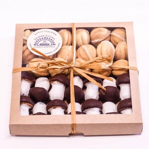 Op de afbeelding: Een bruine kartonnen doos met een transparant plastic venster, gevuld met twee soorten koekjes. De bovenste laag bevat ronde koekjes in lichtbruine kleur met een witte vulling. De onderste laag bevat paddenstoelvormige koekjes met een witte bovenkant en een donkerbruine onderkant.