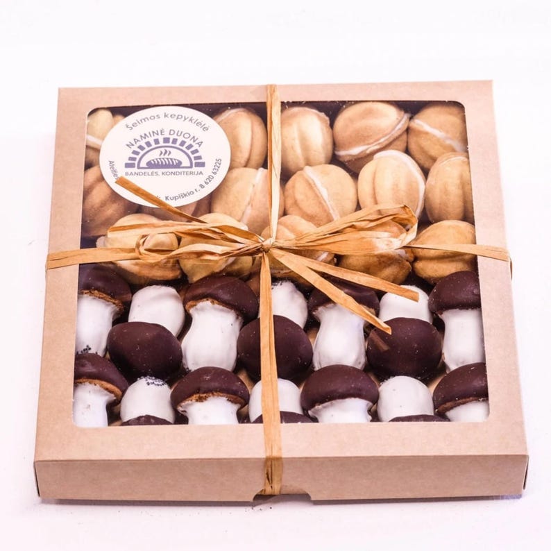 Op de afbeelding: Een bruine kartonnen doos met een transparant plastic venster, gevuld met twee soorten koekjes. De bovenste laag bevat ronde koekjes in lichtbruine kleur met een witte vulling. De onderste laag bevat paddenstoelvormige koekjes met een witte bovenkant en een donkerbruine onderkant.