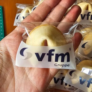 Puede incluir: Una galleta de la fortuna de color beige en un envoltorio de pl&aacute;stico transparente con el texto azul oscuro vfm Gruppe y un logotipo de media luna estilizado. La galleta tiene forma de media luna con el centro plegado.